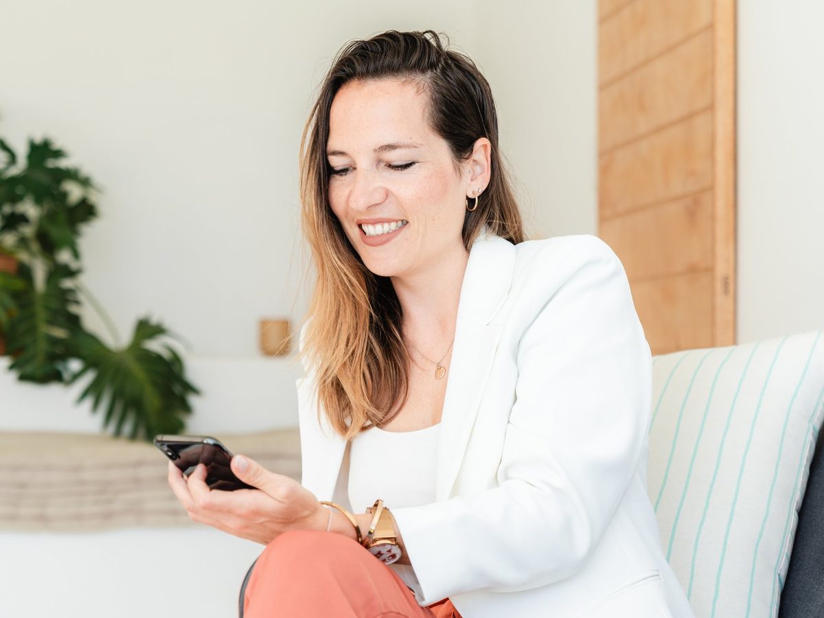 Saskia Drewicke im weißen Blazer mit Smartphone auf einem Sofa